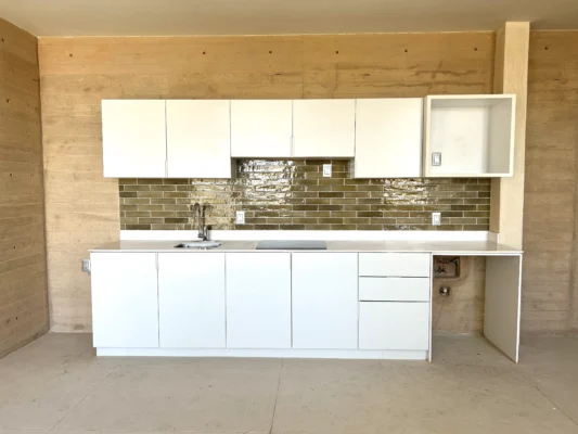 Kitchen with white cabinets and a green tile backsplash. The countertops are a light gray color, and there is a stainless steel sink in the foreground. The faucet is also stainless steel.